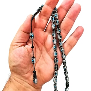 Peut inclure: Un chapelet composé de perles cylindriques noires ornées de motifs à pois blancs et d'accents turquoise centraux. Les perles sont enfilées sur un cordon noir, se terminant par une pièce décorative avec des détails noirs, blancs et turquoise assortis.