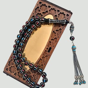 May include: A string of prayer beads featuring black spherical beads with white, red, and blue dot patterns, connected by a silver-toned chain with a multi-strand tassel. The beads rest on a rectangular wooden box with intricate geometric cutouts.