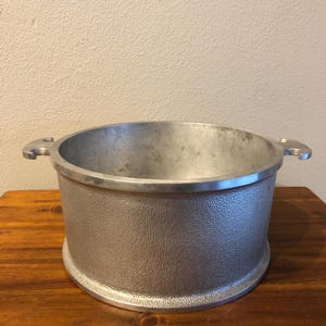 May include: A silver-colored, cylindrical cooking pot with a textured exterior and two side handles. The pot has a wide opening and sits on a slightly raised base. The pot is made of metal.
