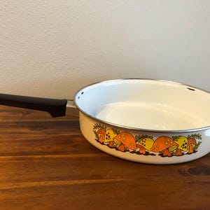 May include: White enamel skillet with a black handle and a decorative band of orange and yellow mushrooms. The skillet has a silver rim and is set on a wooden surface.