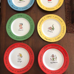 May include: Six decorative plates with zodiac signs. Each plate has a white center with a zodiac illustration and name. The rims are colored blue, yellow, green, yellow, red, and red. The plates are arranged on a brown surface.