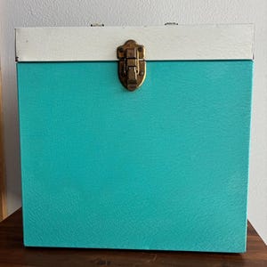 May include: A square storage box featuring a textured turquoise body and a smooth white lid. The box is secured with a central brass-toned metal latch mechanism positioned on the front, connecting the lid to the main body of the container.