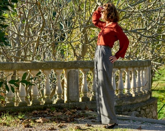 May include: A woman wearing a rust-colored blouse and gray wide-leg pants stands in front of a stone railing. She is looking off to the side, with one hand on her hip and the other hand shading her eyes from the sun.
