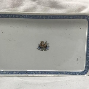 May include: Rectangular white ceramic tray with a blue patterned border. The tray features a central emblem of a ship with the text "REMBRANDT HUDSON" below it. The border has a repeating floral design.
