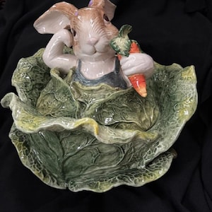 May include: A ceramic figurine of a rabbit holding a carrot, nestled in a cabbage-shaped bowl. The rabbit has brown and white fur, wearing a blue outfit. The cabbage is green with detailed leaf textures.