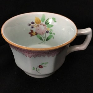 May include: A white teacup with a floral design. The cup has a pink and yellow floral arrangement with green leaves. The rim is trimmed with a gold band and a purple patterned border. A small floral design is on the side of the cup.
