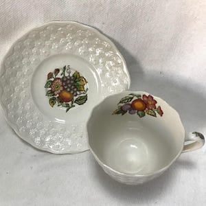 May include: A white teacup and saucer with a floral design featuring fruit and flowers. The saucer has a textured surface.