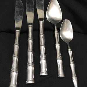 May include: A set of five silver-colored utensils, including three knives and two spoons. The handles have a bamboo-like design. The utensils are arranged on a dark background.