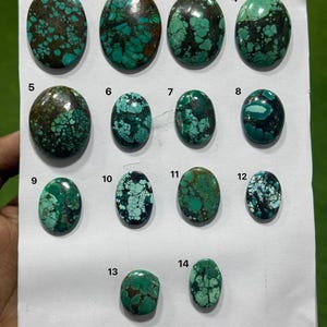 Pode incluir: Uma coleção de catorze cabochões de turquesa ovais polidos dispostos numa folha branca, cada um numerado de 1 a 14. As pedras exibem vários tons de verde e azul com padrões de matriz castanho-escuro ou preto e veios naturais.