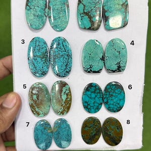 Può includere: Una collezione di sedici cabochon di turchese disposti in otto coppie numerate su un cartoncino bianco. Le pietre presentano varie tonalità di blu e verde con distinti motivi a matrice scura, con forme che variano da ovali a rettangolari.