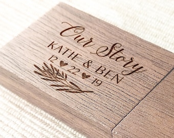 Op de afbeelding: Een houten USB-stick met de inscriptie "Our Story", de namen "Katie & Ben" en de datum "12 22 19". Een takje en hartsymbolen zijn ook gegraveerd. De stick is lichtbruin.