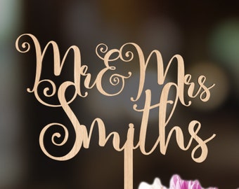 May include: A wooden cake topper with the words "Mr. & Mrs. Smiths" in a cursive font. The topper is placed on a white cake with pink and white flowers.