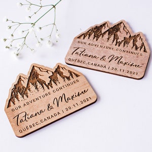 May include: Two wooden wedding favors with a mountain range design. The favors are engraved with the text "OUR ADVENTURE CONTINUES Tatiana & Maxime QUEBEC, CANADA | 20.11.2021".