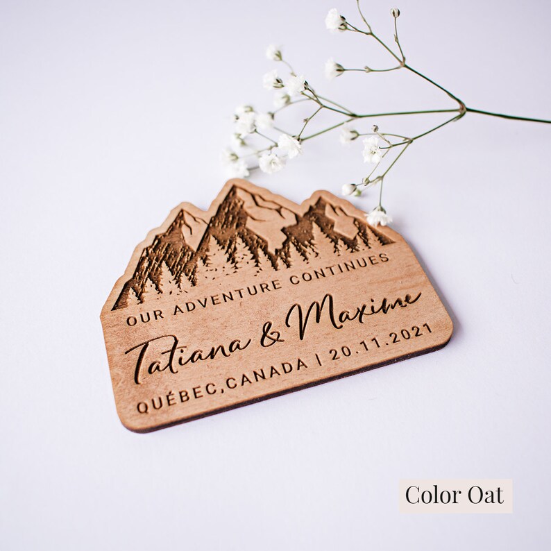 May include: A wooden sign with a mountain range design and the text "Our Adventure Continues Tatiana & Maxime Qu&eacute;bec, Canada | 20.11.2021". The sign is a light brown color and has a natural wood grain.