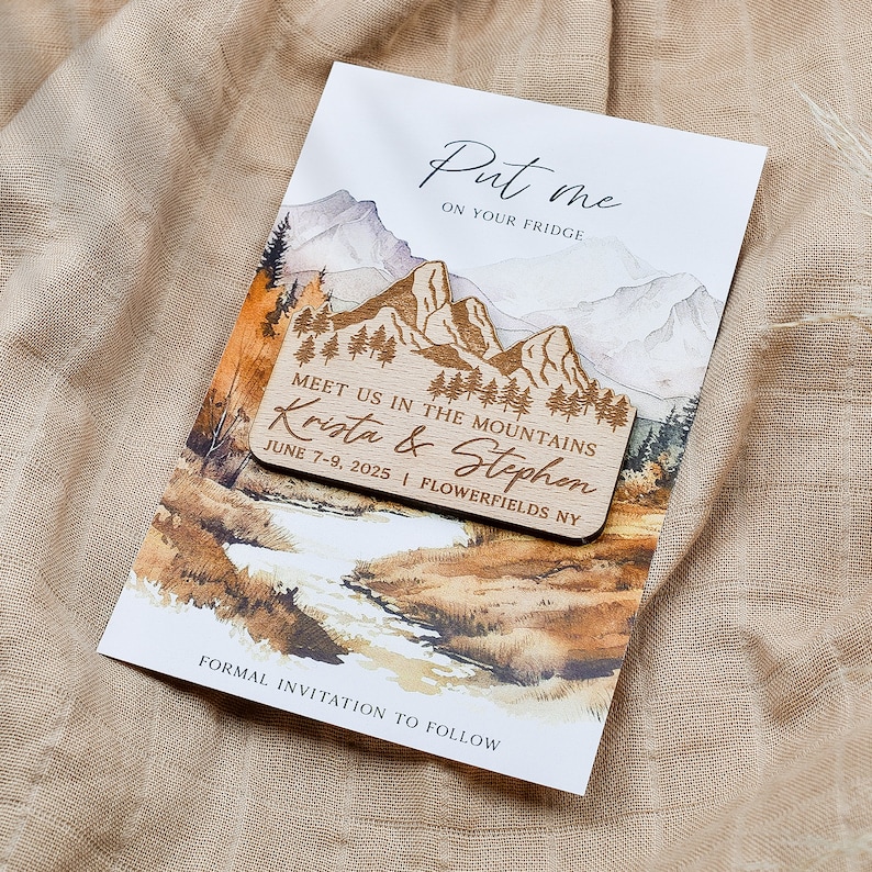 May include: A wooden save-the-date magnet with a watercolor mountain scene. The magnet says "Put me on your fridge" and "Meet us in the mountains Krista & Stephen June 7-9, 2025 | Flowerfields NY".