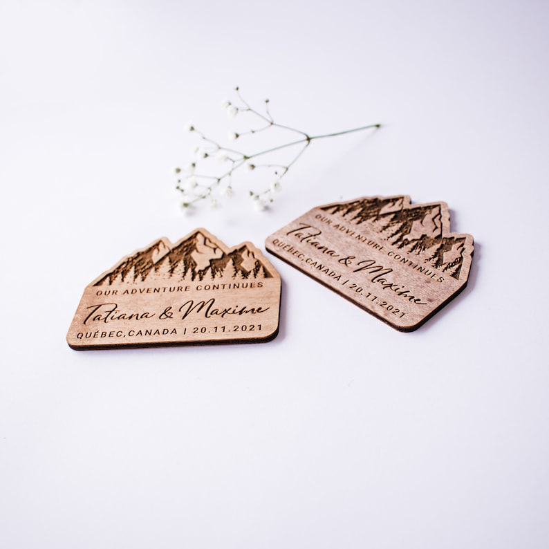 May include: Two wooden wedding favors with a mountain range design. The favors are engraved with the text "Our adventure continues Tatiana & Maxime Quebec, Canada | 20.11.2021".