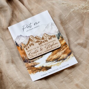 May include: A wooden save-the-date magnet with a watercolor illustration of mountains, trees, and a river. The magnet says "Put me on your fridge" and "Meet us in the mountains Krista & Stephen June 7-9, 2025 | Flowerfields NY".