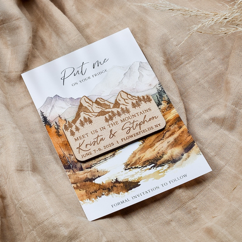May include: A wooden save-the-date magnet with a watercolor illustration of mountains, trees, and a river. The magnet says "Put me on your fridge" and "Meet us in the mountains Krista & Stephen June 7-9, 2025 | Flowerfields NY".