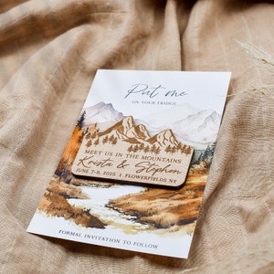 May include: Wooden save the date card with a watercolor mountain scene. The card says "Put me on your fridge" and "Meet us in the mountains Krista & Stephon June 7-9, 2025 Flowerfields NY".