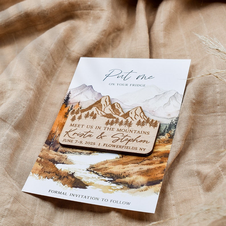 May include: Wooden save the date card with a watercolor mountain scene. The card says "Put me on your fridge" and "Meet us in the mountains Krista & Stephon June 7-9, 2025 Flowerfields NY".