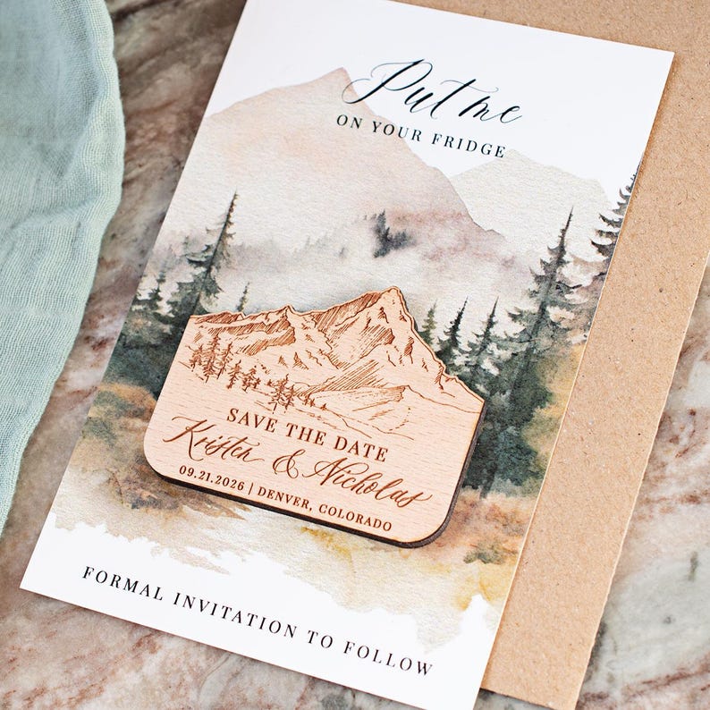 May include: A wooden magnet with a mountain design and the text "SAVE THE DATE Kristen & Nicholas 09.21.2026 | DENVER, COLORADO" is placed on a card with a watercolor mountain scene. The card reads "Put me on your fridge" and "FORMAL INVITATION TO FOLLOW".