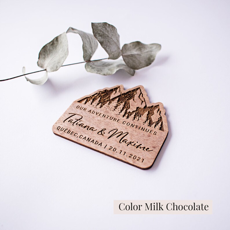 May include: Wooden wedding favor with a mountain range design and the text "Our adventure continues Tatiana & Maxime Quebec, Canada | 20.11.2021". The favor is a light brown color.