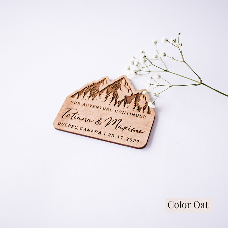 May include: A wooden wedding favor with a mountain range design and the text "Our Adventure Continues Tatiana & Maxime Quebec, Canada | 20.11.2021". The favor is in the color oat.
