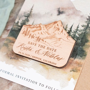 May include: A wooden save-the-date magnet with a mountain and forest design. The magnet is engraved with "Save the Date" and the names "Krista & Nicholas", along with the date and location.