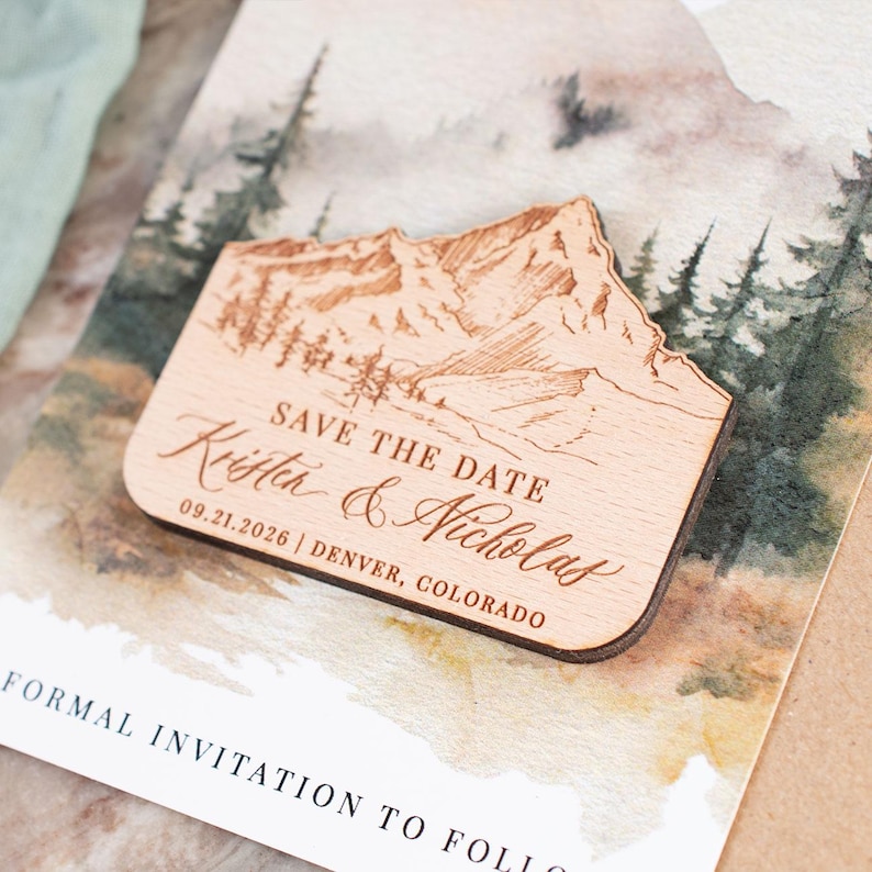 May include: A wooden save-the-date magnet with a mountain and forest design. The magnet is engraved with "Save the Date" and the names "Krista & Nicholas", along with the date and location.