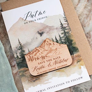 May include: Wooden mountain-shaped magnet with engraved details, featuring the text "Save the Date" and names. The magnet rests on a watercolor-style invitation card with mountain and forest imagery. The card reads "Put me on your fridge."