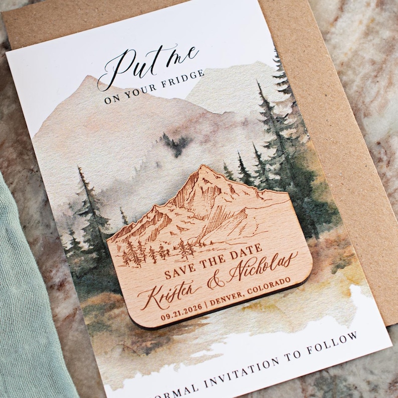 May include: Wooden mountain-shaped magnet with engraved details, featuring the text "Save the Date" and names. The magnet rests on a watercolor-style invitation card with mountain and forest imagery. The card reads "Put me on your fridge."