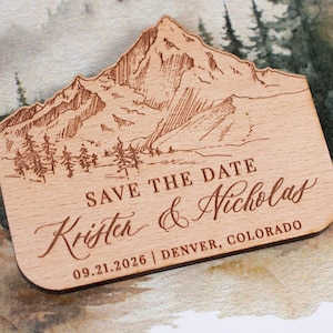 May include: A wooden save-the-date magnet with a mountain and forest design. The text reads "SAVE THE DATE" and the names "Kristen & Nicholas" with the date "09.21.2026" and location "DENVER, COLORADO".