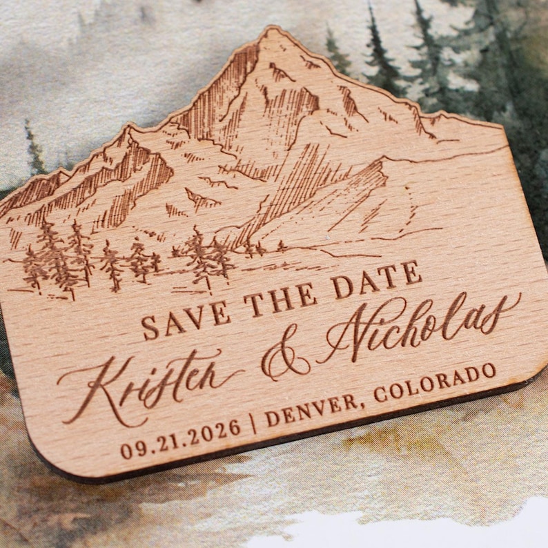 May include: A wooden save-the-date magnet with a mountain and forest design. The text reads "SAVE THE DATE" and the names "Kristen & Nicholas" with the date "09.21.2026" and location "DENVER, COLORADO".