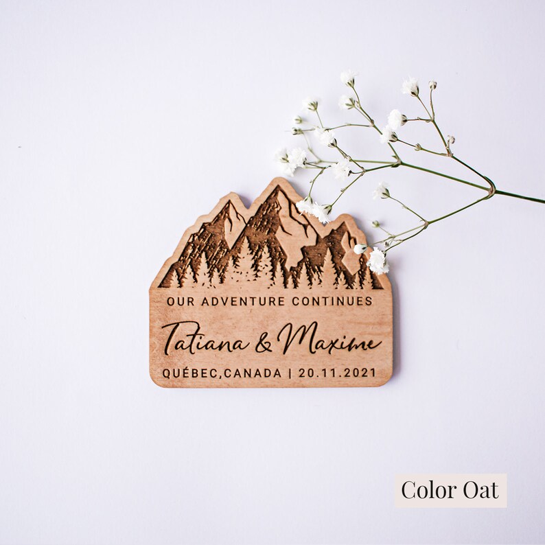 May include: A wooden wedding sign with a mountain range design and the text "Our adventure continues Tatiana & Maxime Qu&eacute;bec, Canada | 20.11.2021". The sign is a light brown color and has a natural wood grain.