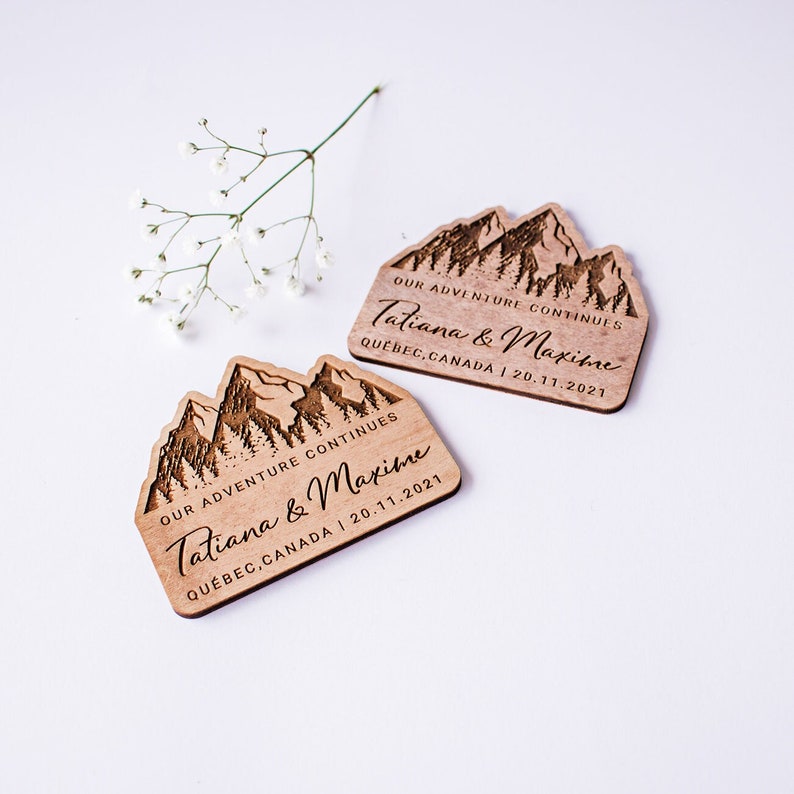 May include: Two wooden wedding favors with a mountain range design. The favors are engraved with the text "OUR ADVENTURE CONTINUES Tatiana & Maxime QUEBEC, CANADA | 20.11.2021".