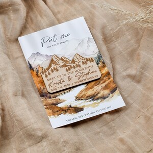 May include: A wooden save-the-date magnet with a watercolor mountain scene. The magnet says "Put me on your fridge" and "Meet us in the mountains Krista & Stephen June 7-9, 2025 | Flowerfields NY".