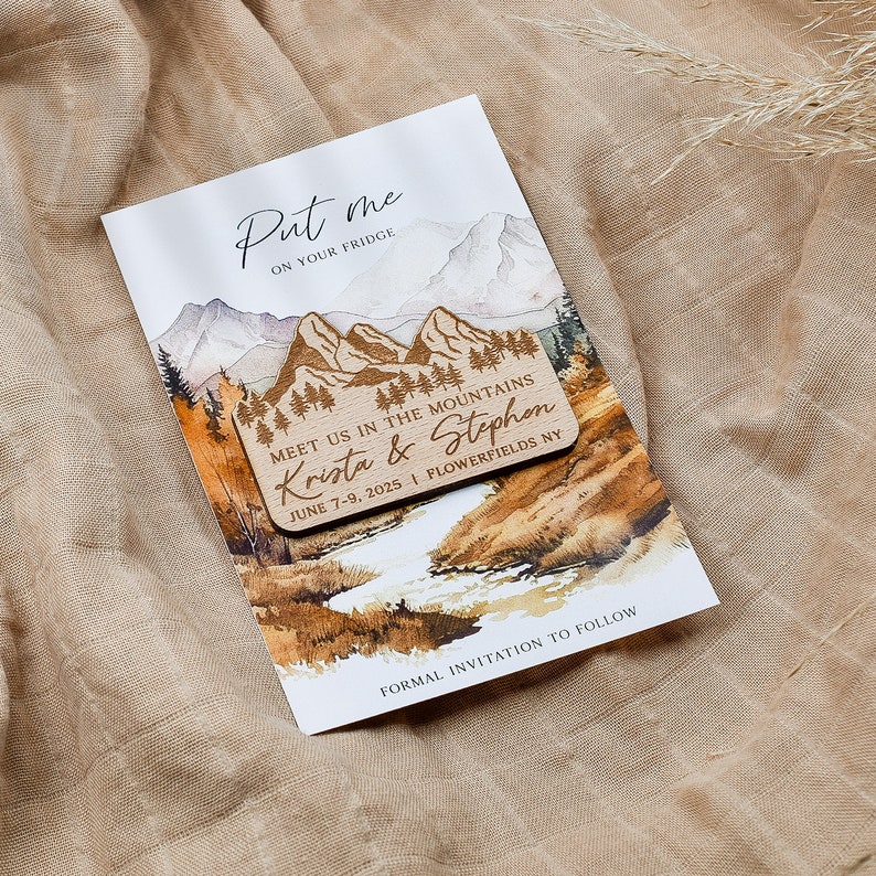May include: A wooden save-the-date magnet with a watercolor mountain scene. The magnet says "Put me on your fridge" and "Meet us in the mountains Krista & Stephen June 7-9, 2025 | Flowerfields NY".