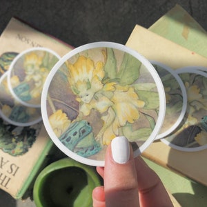 May include: A set of four round stickers featuring a watercolor illustration of a fairy with yellow and green wings, sitting on a green frog. The fairy is surrounded by yellow flowers.