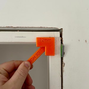 May include: An orange plastic tool with the text "12mm" and "NGH 3D" printed on it. The tool is being used to measure a gap between two pieces of white trim.