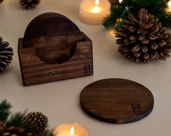 May include: Set of six dark brown wooden coasters with a holder. The coasters are round and have a letter "H" engraved on them. The holder is also dark brown wood and has a letter "H" engraved on the front.