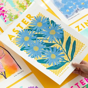 May include: A botanical art print showcasing blue aster flowers with yellow centres and green leaves, set against a yellow and white chequered background. The print is labelled "ASTER" and includes the text "YVE BOTANIC CO." and "L.A. Nursery".