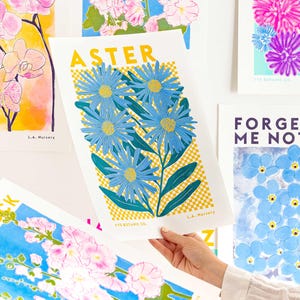 May include: A selection of floral art prints showcasing diverse flowers. One print features the word "ASTER" above blue aster flowers with yellow centres and green leaves. Other prints include orchids, forget-me-nots, and other colourful floral designs.