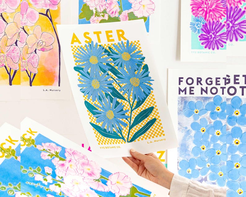 May include: A selection of floral art prints showcasing diverse flowers. One print features the word "ASTER" above blue aster flowers with yellow centres and green leaves. Other prints include orchids, forget-me-nots, and other colourful floral designs.