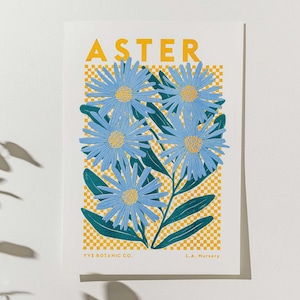 May include: A botanical art print featuring blue aster flowers with yellow centres and green leaves. The word "ASTER" is printed in yellow at the top. The background has a yellow and white checkered pattern. The print is from YVE BOTANIC CO. and L.A. Nursery.