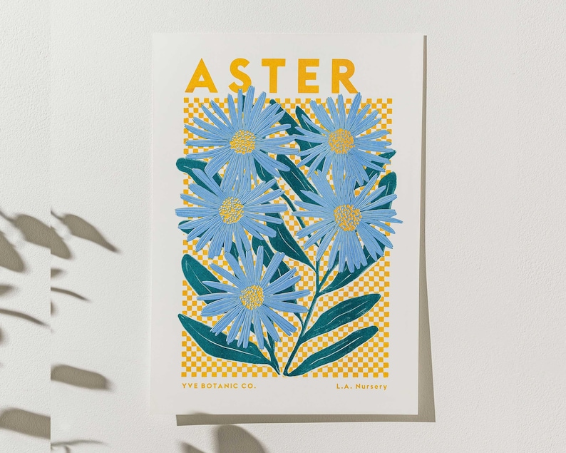 May include: A botanical art print featuring blue aster flowers with yellow centres and green leaves. The word "ASTER" is printed in yellow at the top. The background has a yellow and white checkered pattern. The print is from YVE BOTANIC CO. and L.A. Nursery.