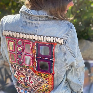 Könnte beinhalten: Hellblaue Jeansjacke mit bunten gestickten Patches und Spiegelakzenten auf dem Rücken. Die Jacke hat eine Reihe weißer Muscheln am oberen Rand. Die Patches enthalten rote, violette und gelbe Designs.