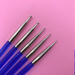 May include: Five nail art dotting tools with silver metal tips and blue handles. Each tool has a different sized ball at the end. The tools are arranged diagonally against a pink background.