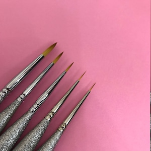 May include: Five silver and gold detail nail art brushes with different sized tips on a pink background.