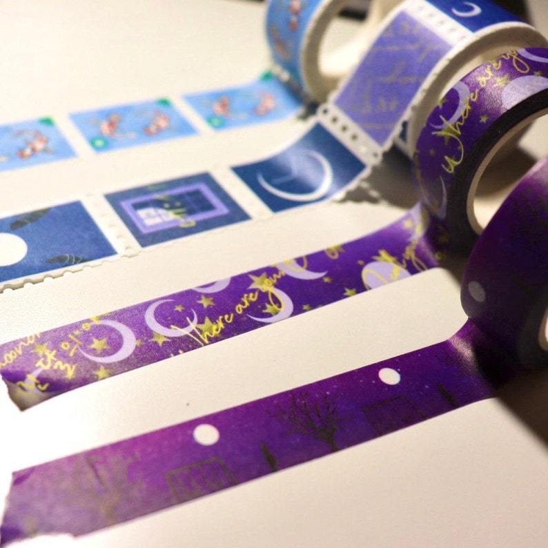 May include: Assortment of decorative washi tapes with moon and star patterns in shades of blue and purple. The tapes feature gold accents and the text "There are stars..." on one of the tapes.
