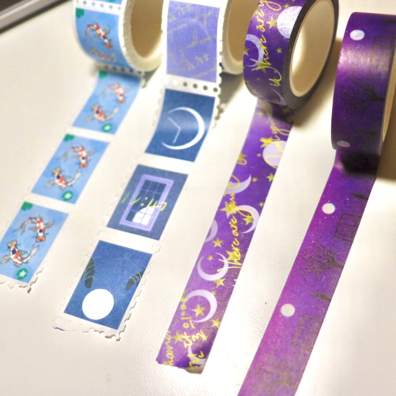 May include: Four rolls of decorative washi tape with different designs. The first roll has a blue background with white and gold koi fish. The second roll has a blue background with white and gold crescent moons and a window with a gold star. The third roll has a purple background with gold stars and crescent moons and the text "Moon over June." The fourth roll has a purple background with gold stars and a silhouette of a tree.